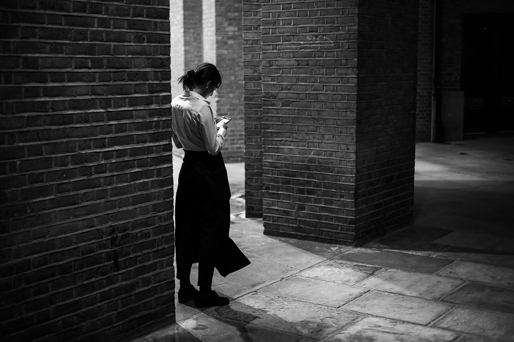 Black and white street photo of a woman checking her phone between brick archways
